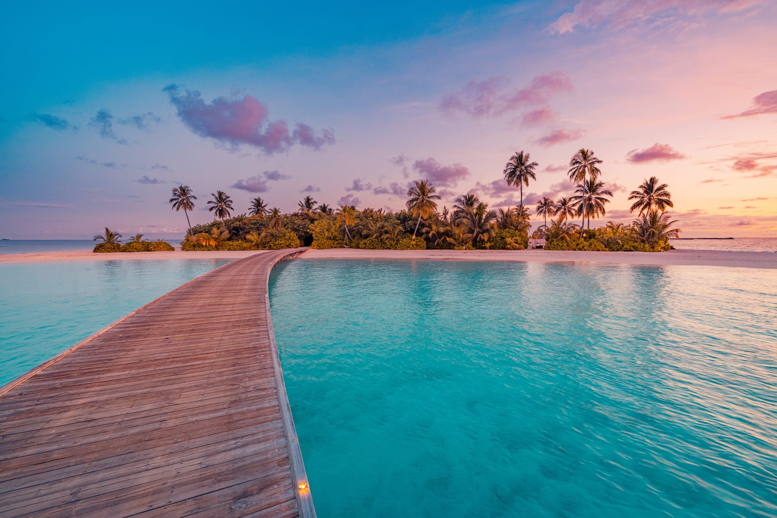 Wooden jetty leading to a palm-fringed island at dusk